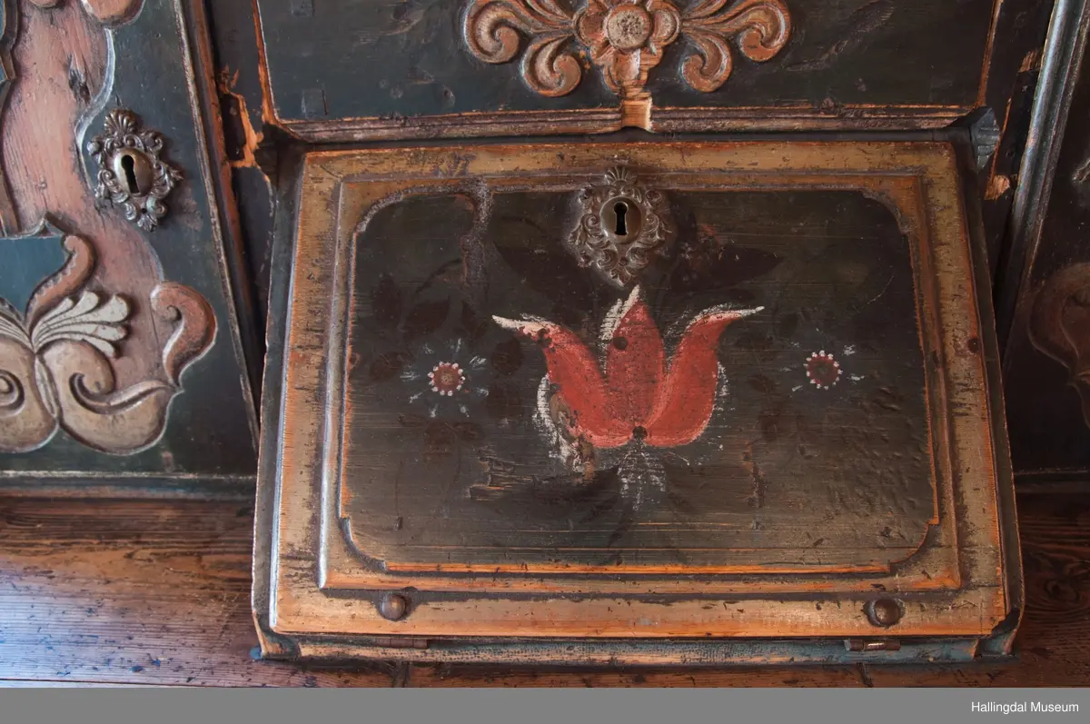 Delene satt sammen v.hj.a. tapping og trenagler.  Gavlen buede sider med skåret kronlist, delt i 3 profilerte, med opprullede ender.  Kronlister fortsetter på sidestykkene.  I toppen av kronlist hjerteformet, gj.brutt utskjæring opp ned.  Gavlens sentralfelt utskjæring i dyp flatskurd.  To løver, vendt mot hverandre, griper om hellebordskaft.  En stake som øverst kløyves i to bjørnehoder i profil skiller løvene.  Gavlen (el. baldakinens) forplate danner fremsiden i skuff i skapets bredde og dybde.  Overskapet:  Åpen hylle øverst, ca. 25 cm høy.  Under hyllen er det delt i 3 felt.  Ett skap i hvert sidefelt.  Midtfeltet har 2 skuffer adskilt av åpen hylle.  H. Skuffe: 12,5 cm.  H. åpent rom: 17 cm.  Under nederste skuff, skap med skråsider som stikker ca. 15 cm ut fra forveggen på overdelen.  Skapet dekkes av liggende skråklaff.  Festet jrenbeslag og hengsler nederst.  
En stor skuff i hele skapets bredde, 6 cm høy nederst, 32 cm dyp.  Over skuffen er rommet delt i 3 felt.  Hvert sidefelt har 2 skuffer og midfeltet en dyp skuff.  Midskuff har fylling med utskåret medaljong, klaff og hylle og skuffen rødmalt. .................................................