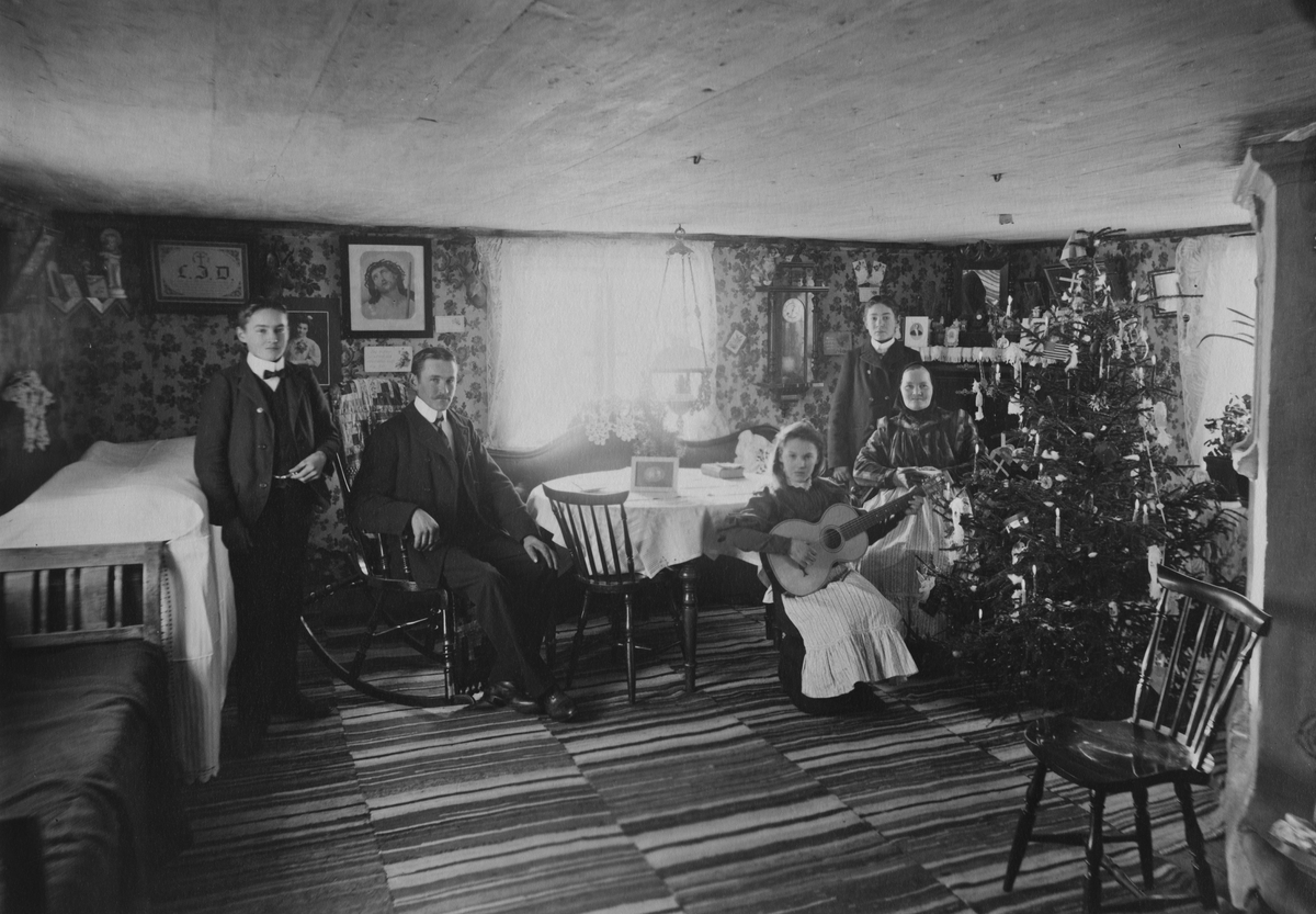 Interiör från en lantgård vid 1900 talets början. Granen är klädd inför julfirandet och en ung kvinna spelar gitarr.