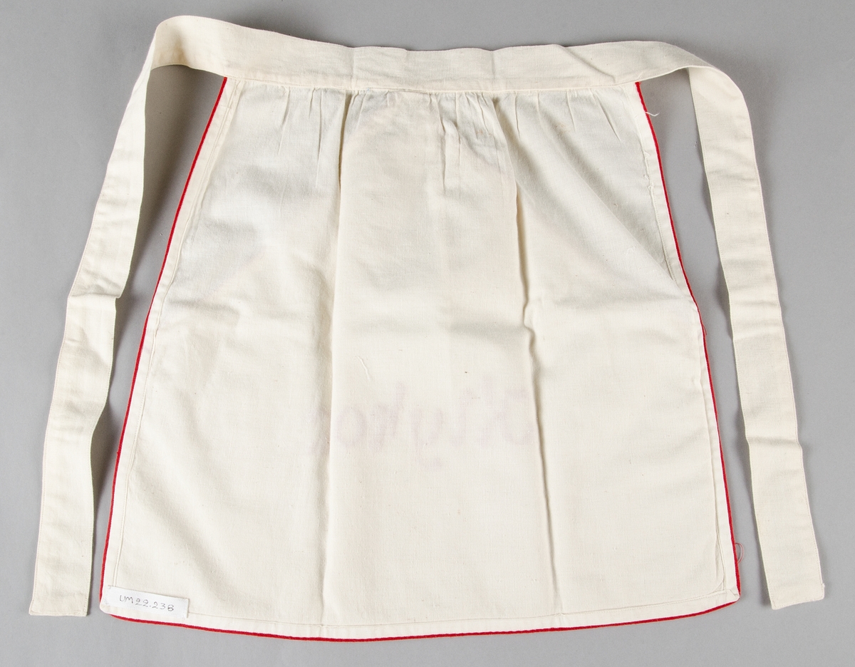 Kort midjeförkläde av vit bomullslärft med röd smal bandkant, med stor ficka med rött plattsömsbroderi: "Klyko". Linning och knytband i samma tyg.