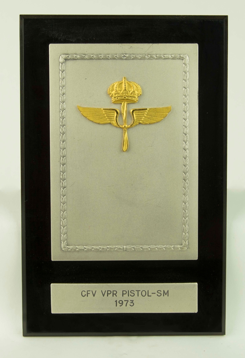 Prisplakett monterad på svart träplatta. Runt plaketten finns en bård av blad. Plaketten har flygvapnets emblem. Under plaketten finns en rektangulär metallplatta med inskriptionen: CFV VPR PISTOL-SM 1973.