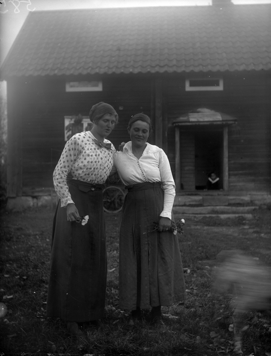 Två kvinnor med blommor i handen står på gårdsplanen, Uppland 1917