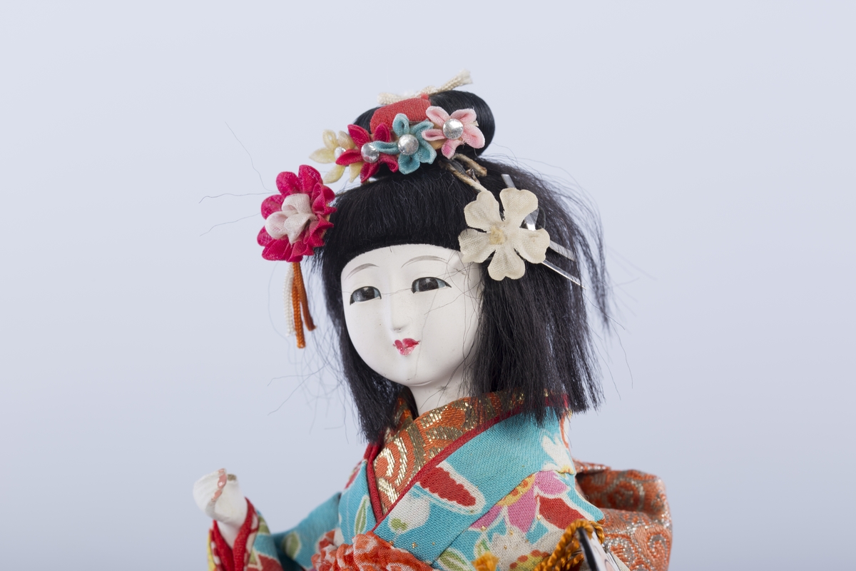 Dukke på lav svartlakkert fot. Dukken er ikledd en kimono og geta (høye tresko), og holder en tromme. Blomster i håret.