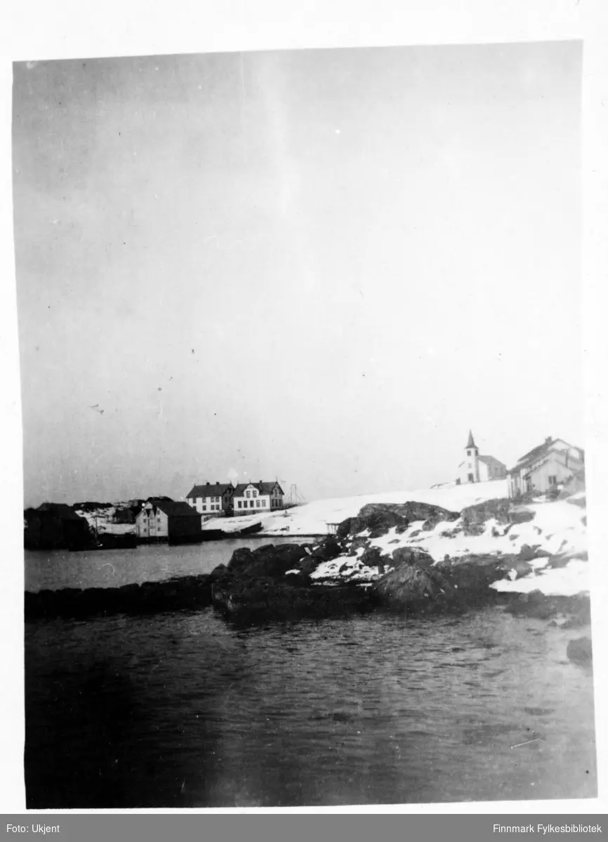 Dette er et postkort fra Øksfjord. Nederst ved sjøen står det boder for fiskemottak. Ovenfor og til venstre står trolig handelsbestyrerens bolig og til høyre for den, et gjestgiveri. Om man går enda lengre til høyre står det en kirke som ble innviet i 1898. I forgrunnen av bildet kan man se fjærasteiner og snø.