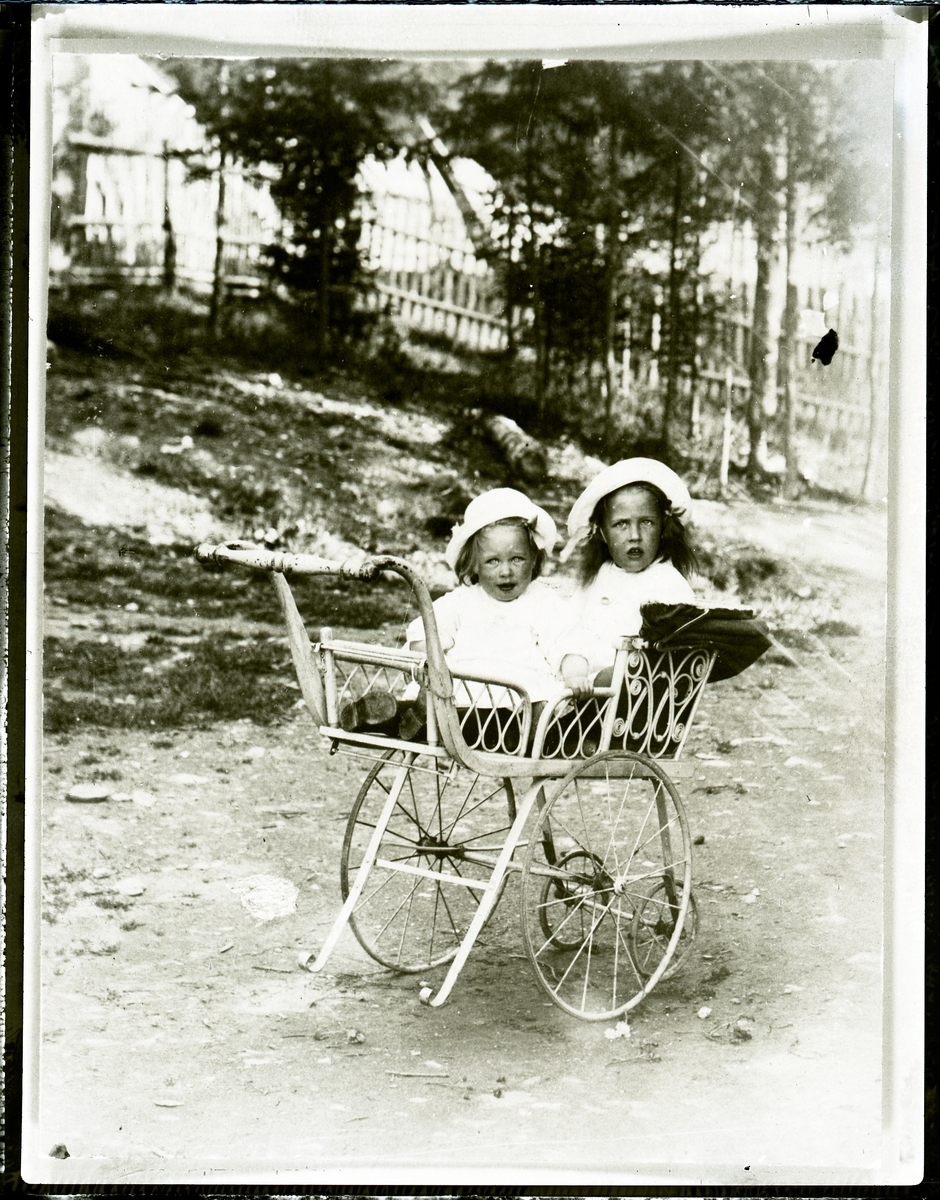 To jenter sittende i barnevogn . Sigrid f. 1915 gift Hall og Synnøve f.1916 gift Kolstad. Bildet er tatt utenfor kjøpmann Melby sin forretning og jentene er hans døtre. Bildet er tatt ca 1917.