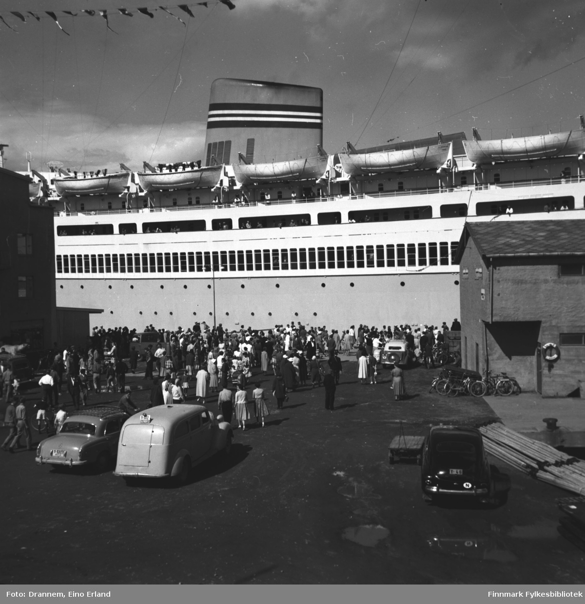 Et stort passasjerskip ligger ved Dampskipskaia i Hammerfest. Mange personer står på kaia mellom FFRs administrasjonsbygg, til venstre, og godsterminalen, til høyre. Dette er Den Norske Amerikalinjes M/S Bergensfjord, bygget i 1956.