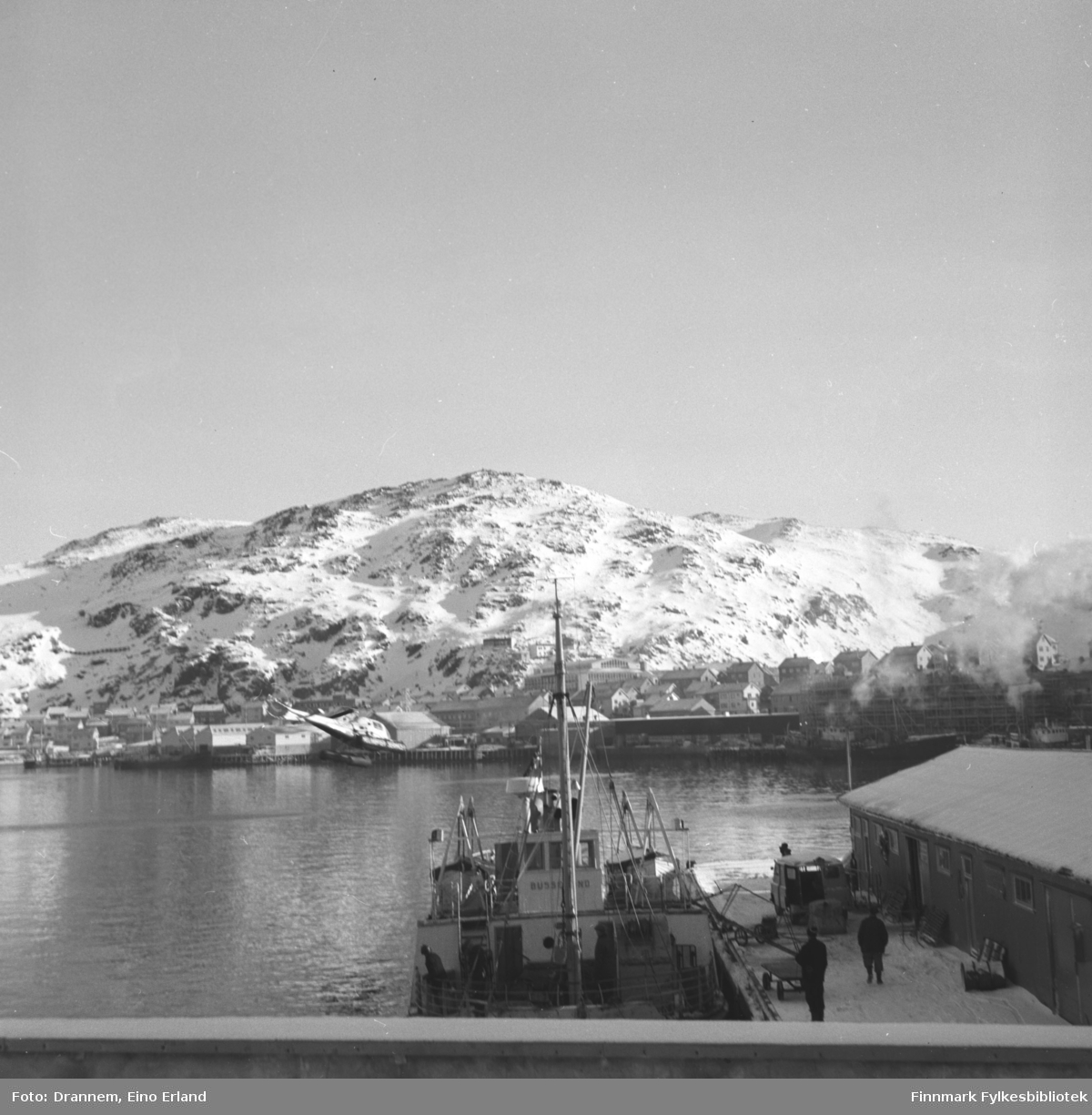 Del av Hammerfest fotografert fra Tollboden med båten "Bussesund" som ligger i forgrunnen ved Dampskipskaia. Mollafjell ses i bakgrunnen. Et helikopter går inn for landing i havneområdet.