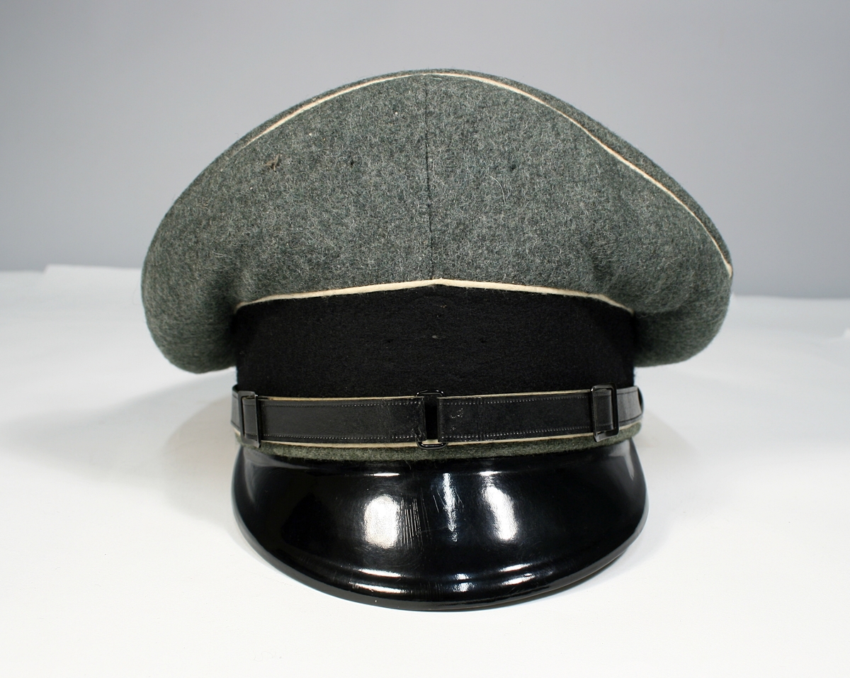 Tysk uniformslue i grågrønt ullstoff med sort luebånd og snor og skjerm i sort lakk. Lyse paspoiler.
