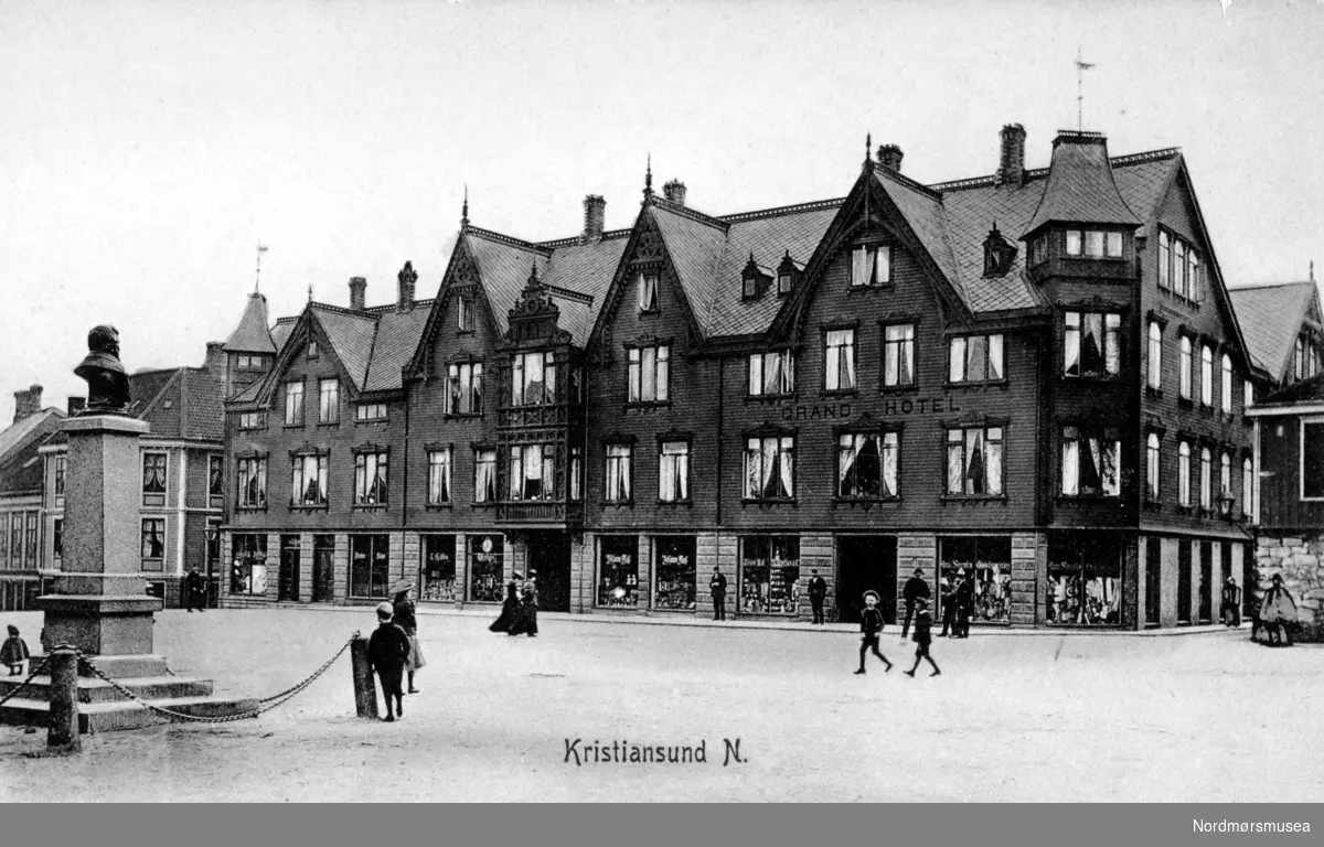 Det første Grand Hotell på Torget på Kirkelandet i Kristiansund, i begynnelsen av 1900 tallet.

Historie: Etter en alvorlig økonomisk krise i 1880 årene var byen nå atter preget av fremgang og optimisme. Byen hadde på denne tid en befolkning på 11 til 12000 innbyggere. Det var nå behov for et nytt standmessig hotell for reiselivsnæringen, foretningsfolk samt byens egen befolkning.

Gustav W. Dahl som var født i Trondheim i 1863, der han i første halvpart av 1890 årene hadde arbeidet som hotelltjener blant annet ved Grand Hotell cafe. At han ville prøve seg på hotelldrift i Kristiansund skyldes nok mye hans kone som var fra Kristiansund, og gjennom henne var blitt kjent med byen. 

Dette ledet til at i første halvpart av 1890-årene ble det oppført et boligkompleks på tomten til den gamle Prestegården, og som hadde fasade mot Torvet og Bernstorffstedet. Det var Stadskonduktør Hagbart Brinchmann som var byggherre og eier av bygget, hvor Grand Hotell etablerte seg i.

Mai 1895 kunne man lese i en annonse i to av byens aviser at byens Grand Hotell åpner dørene 1. juni 1895, og annonsen var signert hotellets eier Gustav W. Dahl. 

Hotellet hadde 30 værelser, og foruten gjesterom hadde det udpakningsværelser, en elegant Salon, Læseværelse og Spisesal.

Driften av hotellet ser ut til å ha godt bra, for i 1897 etablerte Gustav W. Dahl seg som restauratør på Øvre Bakklandet i Trondheim. Dette kjøpet ble fulgt opp av et nytt kjøp våren 1900, da han også kjøpte Trondheims Grand Hotell.

Ettersom Gustav W. Dahl nå hadde satset så hardt i Trondheim ble det behov for en ny innehaver for Grand Hotell i Kristiansund. 20. november 1900 kunne man da også lese i avisen i Kristiansund at nye lederen for hotellet nå var Nils Nilsen.

Nils Nilsen var født i Sand i Ryfylke i 1869. Han hadde vært reisefører og tolk for den britiske godseier og vinkongen Fleetwood Sandeman og gjennom hans reiser gjennom flere europeiske land, hadde fått et inngående kjennskap til hotelldrift. På det tidspunkt han tok over driften av hotellet var han faktor ved Sandemans gård Gulla i Surnadal. Trolig fikk Nils Nilsen også økonomisk støtte fra sin arbeidsgiver til å kjøpe hotellet.

En av forandringene Nils Nilsen gjorde ved hotellet var å installere hustelefon på alle rom.

En av de større begivenhetene hotellet opplevde var å være vertskap for kongefamilien på dens kroningsferd den 18. juni 1906. Nils Nilsen sørget først for en bedre borgermiddag i gymnastikksalen og i andre rom ved Enggatens skole. Hans Majestet Kong Haakon VII overnattet senere på hotellet, mens Hennes Majestet Dronning Maud overnattet på  kongeskipet Heimdal for å få mer ro neste morgen. Morgenen etter ble det arrangert en avskjedsfrokost for kongen, der medlemmer av formannskapet og embetsmenn med fruer deltok. 

Så i 1899 døde Hagbart Brinchmann som eide bygningen der Grand Hotell holdt til, og hans enke, Laura Susanne Brinchmann (født Øwre) solgte da bygningen til Nils Nilsen våren 1907. Nils Nilsen var nå blitt eier av både hotellet og bygningen.

Men så den 8. november 1907 brøt det ut brann i hotellets vedbu. Brannen ble oppdaget av nattevakten, og alarm ble slått. Mer enn 300 mann kom raskt til stede, samt 5 fra sjødampsprøyta, men det ble snart klart at det store trekomplekset ikke kunne reddes. To mennesker mistet livet i brannen. Disse var søstrene Anna og Augusta Sørensen som drev motehandel i første etasje av hotellet. Den flotte trebygningen i utført sveitserstil var nå blitt slukt av flammene.

Ikke lenge etter besluttet Nils Nilsen å bygge et nytt hotell på den samme tomten. Arkitekten ble Kristen Tobias Rivertz, og det nye hotellet som ble oppført i jugendstil, stod så ferdig januar 1909.

Referanse: En opplevelsens festlabyrint av Egil Husby, side 7 - 16. Nordmøre Museums fotosamling.
