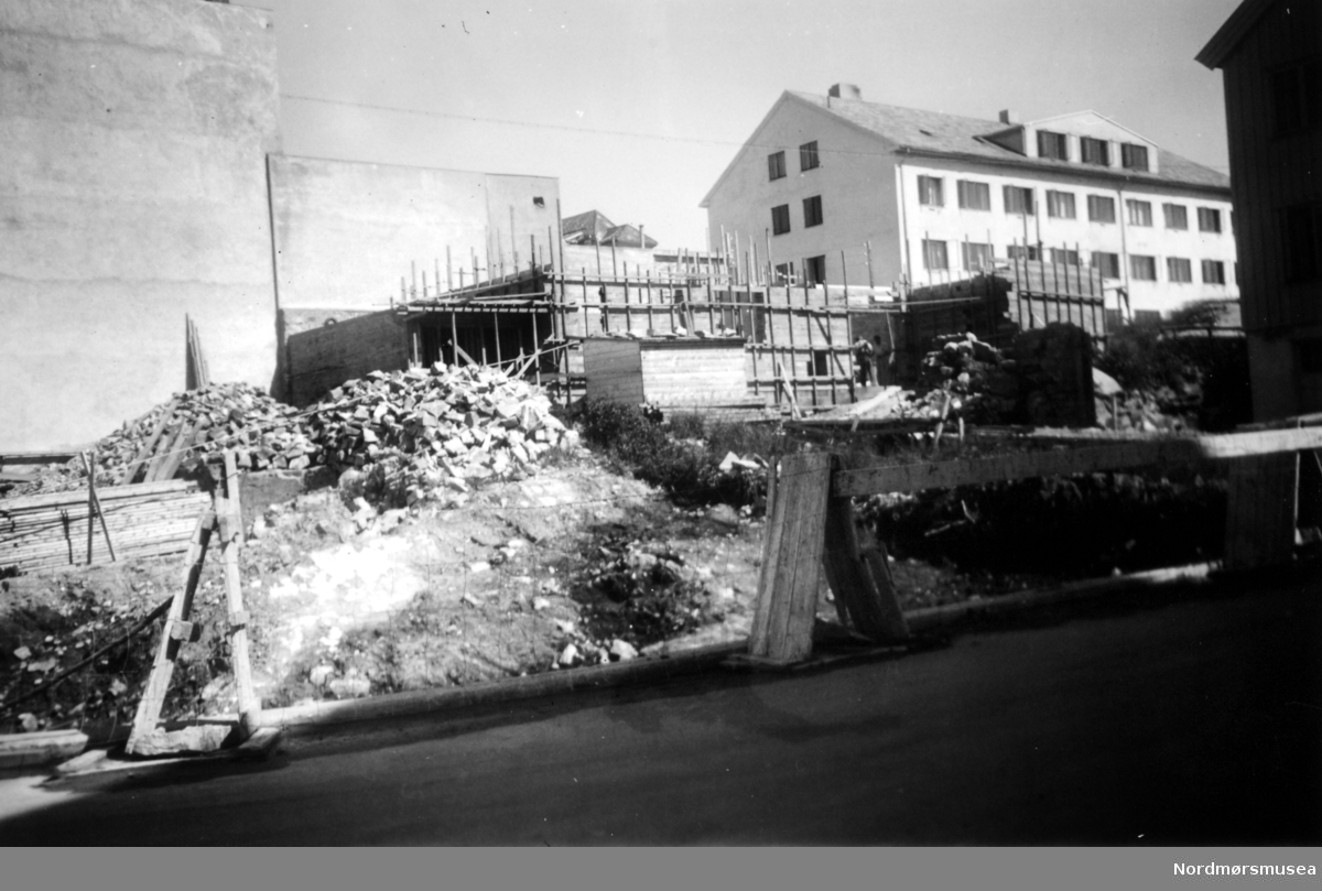 En del av den gamle Metodistkirkens tomt ryddes for Byggmester Hovdes nye gård i Langveien, hvor vi ser hans verkstedbygning som reiser ser på tomten. Ellers så ser vi Lars Hansens konfeksjonsfabrikk i bakgrunnen. Fotografert fra sørvest, trolig på begynnelsen av 1950 tallet. Fra Nordmøre Museums fotosamlinger. 




