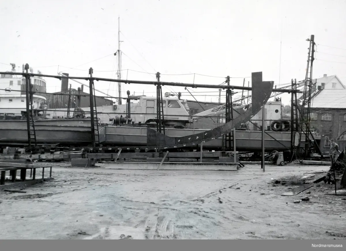 Bildet viser at kjølen og baugsenterplaten er reist på beddingen på B/F”Norddalsfjord” Storviks Mek. Verksted bnr.14 på Dahle ved Kristiansund.
”Norddalsfjord” ble levert til Møre og Romsdal Fylkesbåter 15. mars 1961 og hadde følgende hoveddimensjoner: L 31,20 m x B 8,55 m x D 3,35 m og hadde en tonnasje på 159 bruttoregistertonn. Fremdriftsmaskineriet består av 3 Volvo Penta turboladede dieselmotorer type TMD96 på til sammen 420 hk som via kilremdrift var koblet til et felles gir og propellaksel med vribar propell, slik at hver enkelt av motorene kunne kjøres separat. Fergen hadde 2 Bolinders vekselstrømsaggregater type 1052MG på 23 hk hver tilkoblet en generator på 17 kW. Fergen var utstyrt med elektrohydraulisk styremaskin.
Fergen har plass til 18 personbiler og har sertifikat for 160 passasjerer. Forut er det innredet 6 lugarer for offiserer og restaurantpersonale og akterut en mannskapslugar for 4 personer og toppfarten er 11,4 knop og marsjfarten 10,5 knop.
Ferga er verkstedets første nybygg etter B/F”Trygge” som ble levert i 1938.
Bak nybygget, på patentslippvogna klinkbygd i stål 1918, den største slippen mellom Bergen og Trondheim, ses kranprammen ”Uthavn”, forrest, og mudderfrakteprammen, bakerst, tilhørende Statens Havnevesen.
På såpehellingen fra 1901, bak prammene, ses M/S”Greip” og foran rorhuset til ”Greip” ses støperibygningen.
Helt til høyre i bildet ses kjelverkstedsbygningen, fra 1905 og revet etter 1980, og over den bolighuset til verkstedet fra ca. 1910 som brant på 1980-tallet, hvor familien Paul Storvik bodde.  Bolighuset var et gammelt meieri i tømmer flyttet fra Straumsnesset og bygd opp ca. 1910 og brant på 1980-tallet.
Til venstre for kjelverkstedbygningen og foran verkstedets bolighus ses litt av taket på kontorbygningen fra ca. 1902 og revet og brant på 1970-tallet.
Helt til venstre forrest i bildet ses en bit av sveiseplanet og helt i høyre kant av bildet ses verksmester Christian Larsen med hatt.
Bildet er fra 30/6-1960. (Info: Peter Storvik) ---
"30/6." Foto fra anleggsområdet til Storvik Mekaniske Verksted på Dale, Nordlandet i Kristiansund. Bildet er datert 30.06.1960. Fra Nordmøre Museums fotosamlinger. variant