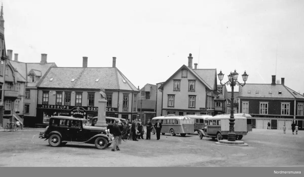 Busser og biler på Torget på Kirkelandet i Kristiansund. Sverdrups bokhandel bak til venstre. Nærmeste bil er en Ford B eller V8 1932, bak den skimtes en Ford V8 1933-34. Ca. tidsangivelse dermed 1935. (Info: Ivar E. Stav - 11.11.2016). Fra Nordmøre Museums fotosamling.