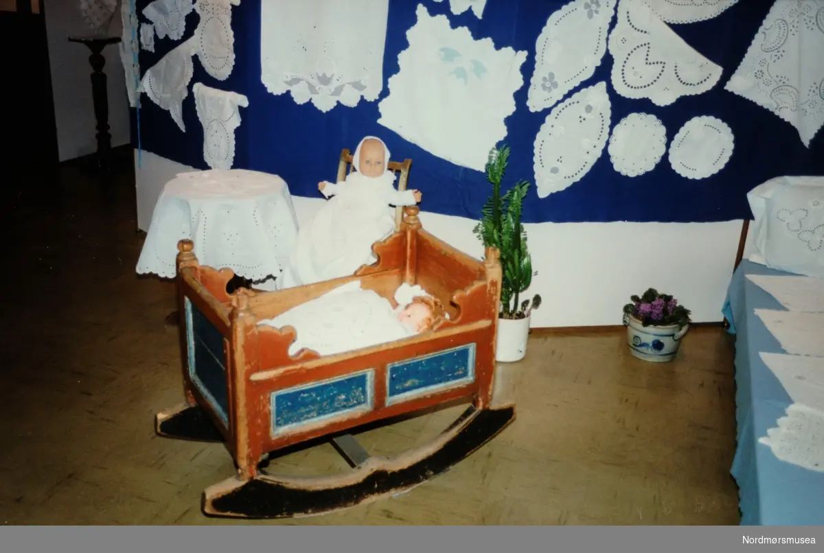 Deler av en utstillingene "Barn" og fylkeshusflidlagets "Opp fra Kistebotnen" som ble holdt i Nordmøre Museums lokaler i Knudtzondalen fra 4. juli 1989. Vugga sies å ha tilhørt Eidsvollsmannen Jonas Rein (som barn i Øksendal). Fra Nordmøre Museums fotosamlinger.