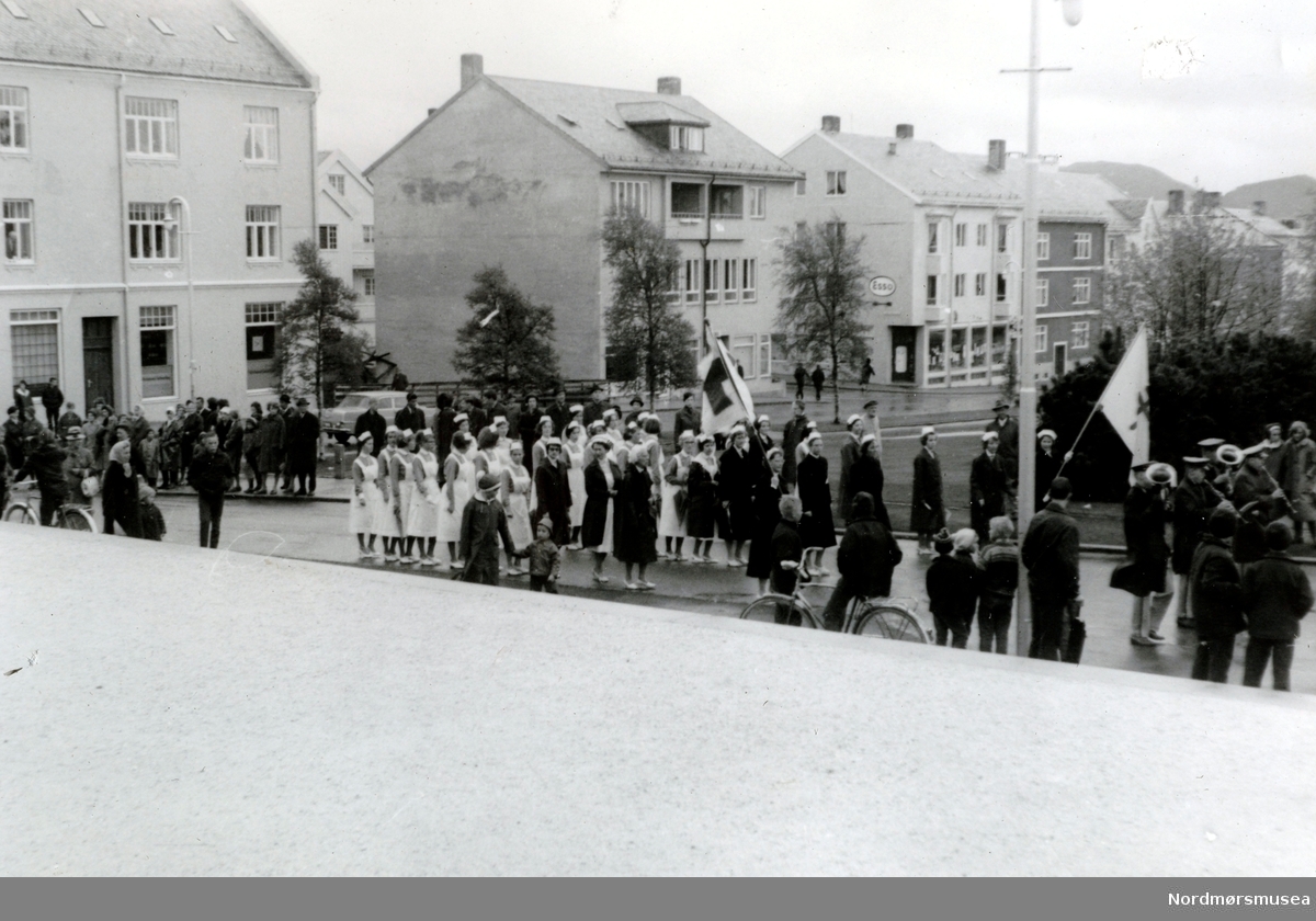 Foto fra et opptog - muligens en 17. mai? - hvor vi ser en gruppe med Røde Kors sykesøstre med faner og musikkorps, foran Kirkelandet kirke i Kristiansund.  Fra Ida M. Knudtzons fotosamlinger, nå en del av Nordmøre Museums fotosamlinger. 
