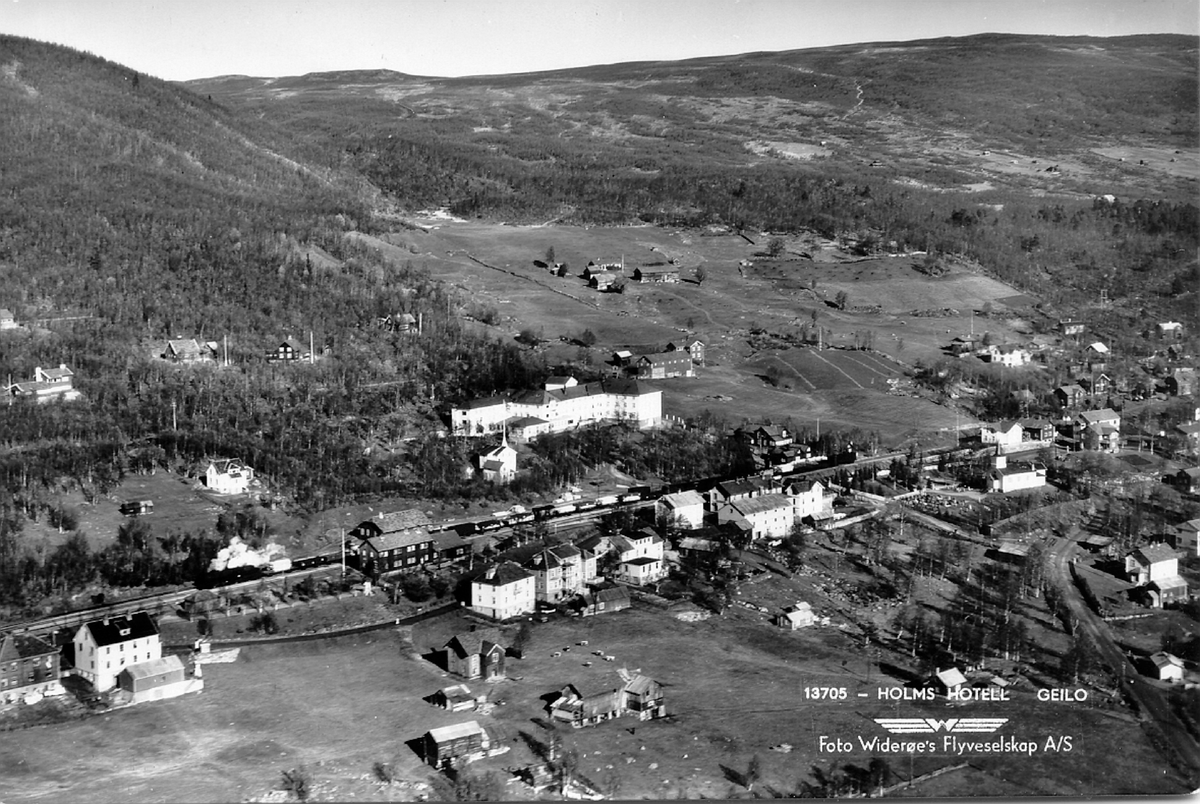 Flyfoto fra Geilo sentrum. Et godstog retning Bergen, trukket av damplokomotiv, står på jernbanestasjonen