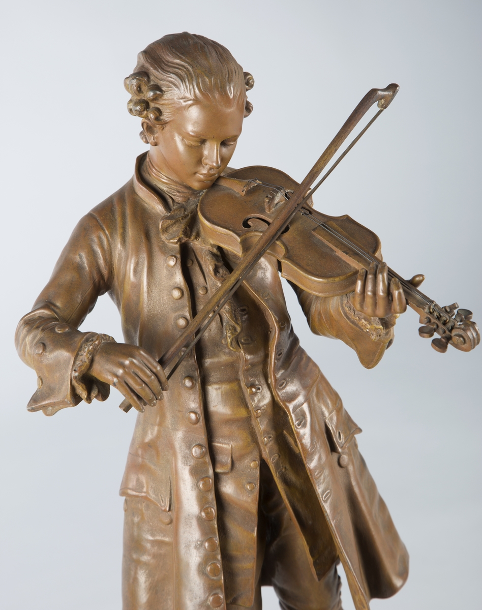 Forestiller ung mann i rokokko-antrekk som spiller fiolin. (Mozart?). Mann står på en bronsesokkel, som igjen er montert på en sokkel av rød-lilla marmor. Under mannens føtter to ark påskrevet noter, dette også i bronse.