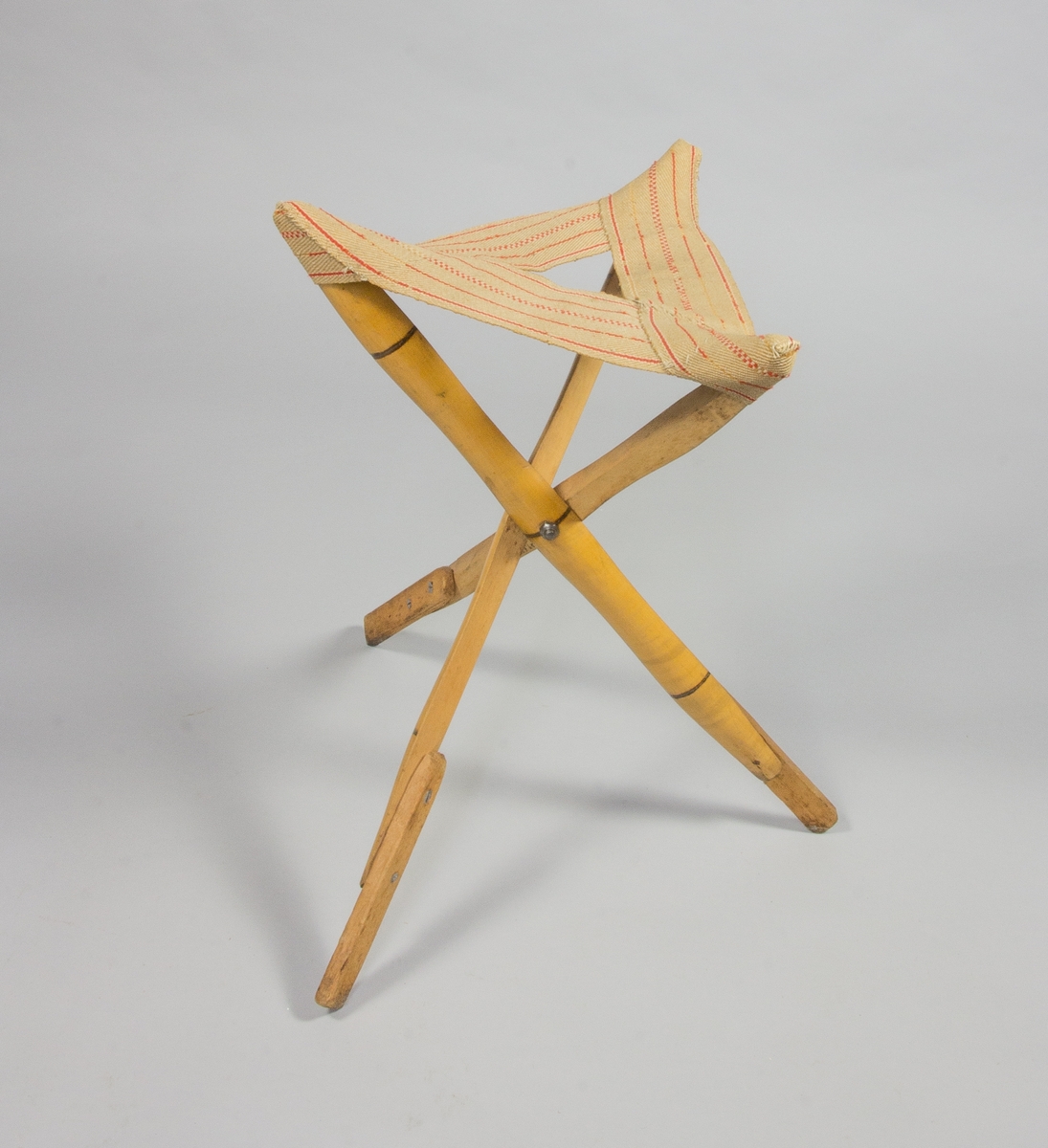 Trebent stol, pall. Hoppfällbar. Ben av bambu, nedtill förstärkta med trä. Trekantig sits av naturfärgad sadelgjord med röda ränder.