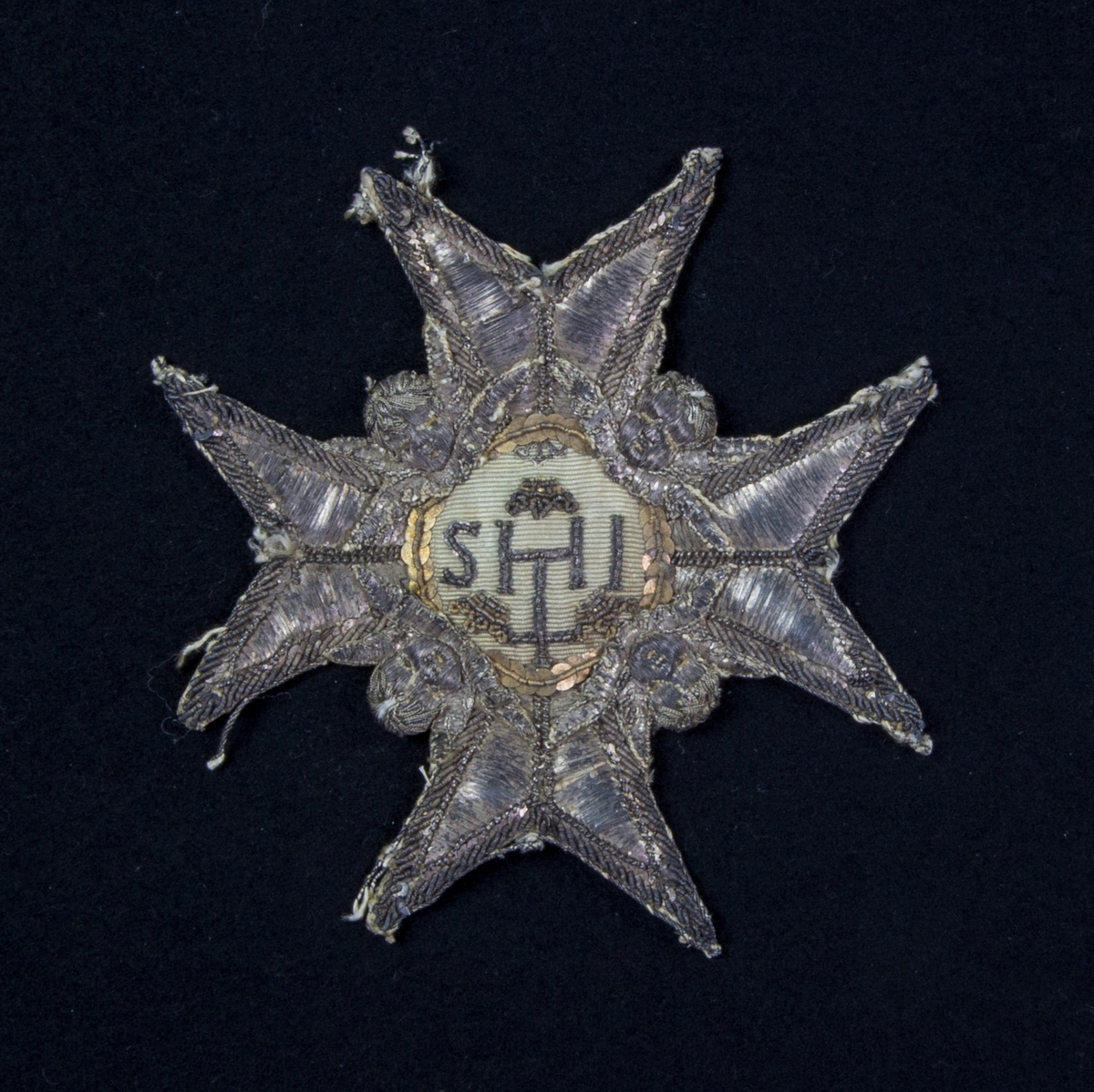 Serafimerordensstjärna med 8 uddar av ljust skinn med broderade ornament av silvertråd, såsom änglahuvuden med mera. I centrum IHS broderat på siden omgivet av paljetter.