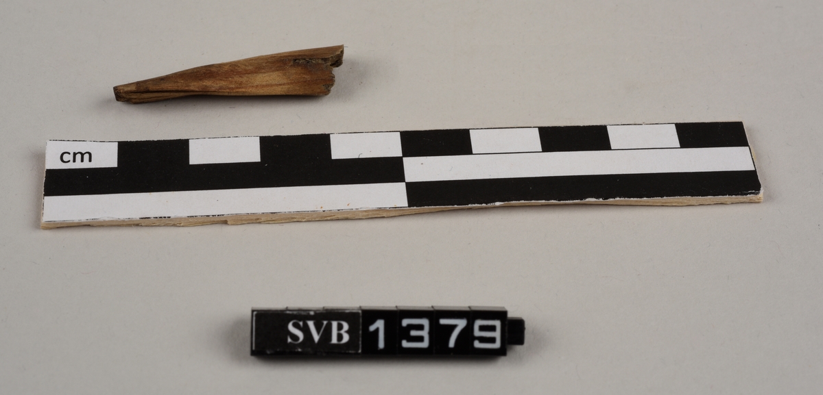 Del av et båtbord. Delen ble saget av. Det er hull til vidje og plugger. Delvis er det rester av både vidje og plugger. Treet fremstår ubehandlet. Det er et materiale som ligner torv/mose mellom bordene.