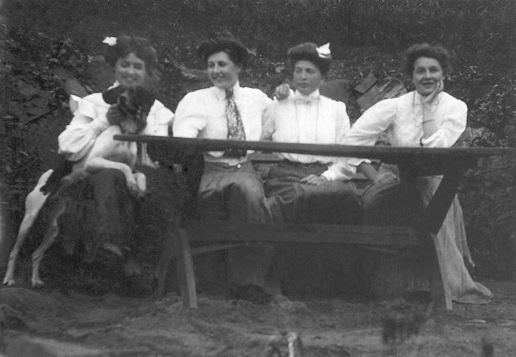 Fyra unga kvinnor i blus och kjol sitter vid ett trädgårdsbord. Kvinnan längst ut till vänster klappar en stövare, som delvis krupit upp i hennes knä.