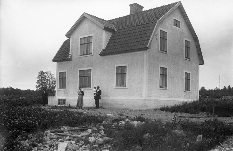 En kvinna och man (Fritz och Agda Bergqvist) som håller i sina barn. De står på baksidan av ett ljust reveterat bostadshus.
Trol. makarna Bergqvists 2:a hem, vid Braås gård, där man bodde från 1921.