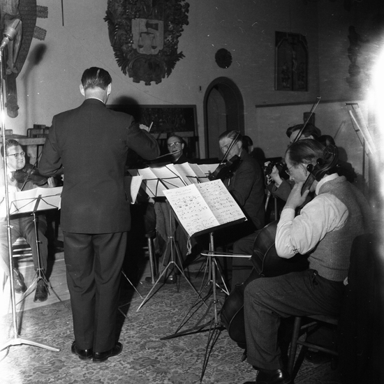 En dirigent dirigerar en musikkår i kyrksalen (numera orgelsalen) på Smålands museum.