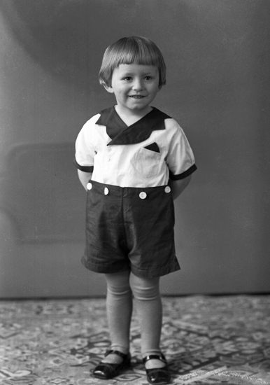 Foto av en liten pojke i mörka kortbyxor och vit tröja, som poserar för fotografen.
Helfigur. Ateljéfoto.