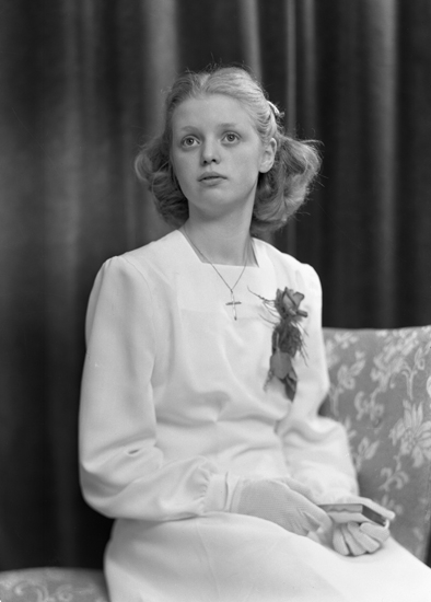 Foto av en ung kvinna i vit konfirmationsklänning. Hon sitter på en stoppad stol.
Knäbild. Ateljéfoto.