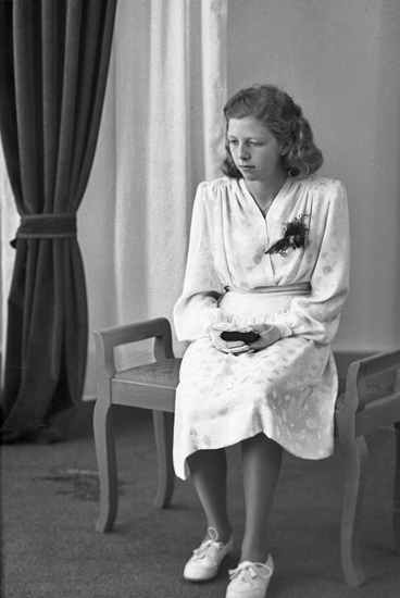 Foto av en ung kvinna i vit konfirmationsklänning. Hon sitter på en stoppad bänk och håller en liten psalmbok i händerna.
Helfigur. Ateljéfoto.
