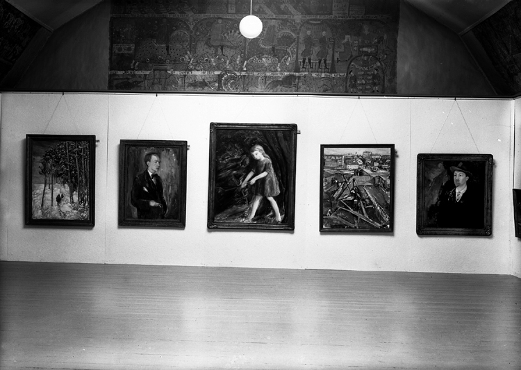 En tavelutställning i Smålands museum, i dåv. textilkammaren.
På väggen hänger ett antal tavlor och överst i mitten syns en målad bonad.