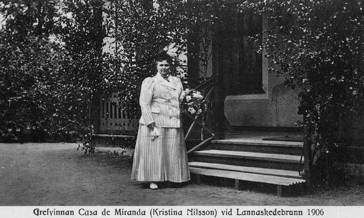 Porträttfoto av Christina Nilsson, vid Lannaskede brunn 1906. Hon står utanför ett hus med veranda. 
Hon är klädd i promenaddräkt.