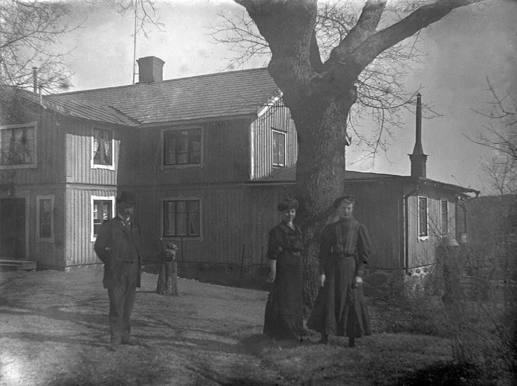 Foto av två kvinnor och en man utanför ett brädpanelat, större bostadshus. (Dansjö Stöfvagård). 

Något suddigt foto.