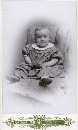 Porträtt (helfigur) av en liten flicka i koltliknande, prickig klänning med stor vit spetskrage. Hon sitter på en tunn fårskinnsfäll i en stol (?) med tofsar, utan armstöd.