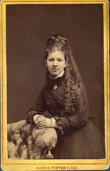 Porträtt (knäbild, en face) av en okänd ung kvinna med hög, mörk frisyr och mörk klänning med hög sammetskrage. Vid kragen syns en medaljong. Hon lutar händerna mot ryggstödet på en stoppad, blommig fåtölj.