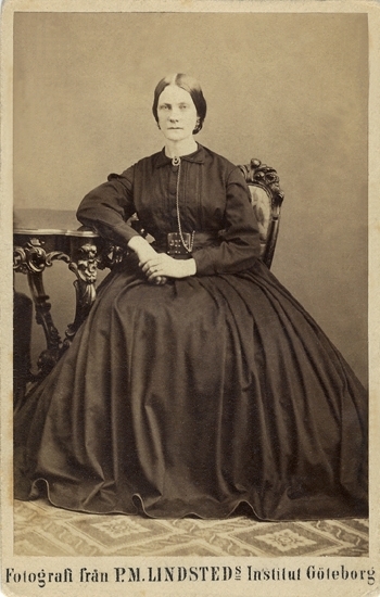 Porträtt (helfigur, en face) av en okänd sittande kvinna i enkel, mörk krinolin. Vid kragen är fästad en klockkedja, som löper ner mot ett skärp.
Hon sitter på en stol i nyrokoko och stöder ena armen mot ett utsirat bord.