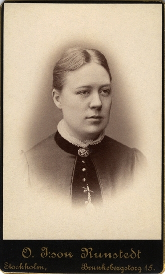Porträtt (bröstbild, halvprofil) av en kvinna i mörk klänning med mörka sammetsdetaljer och hög sammetskrage med vitt krås. 
Vid kragen syns en rund brosch. Längre ner syns början på en klockkedja.