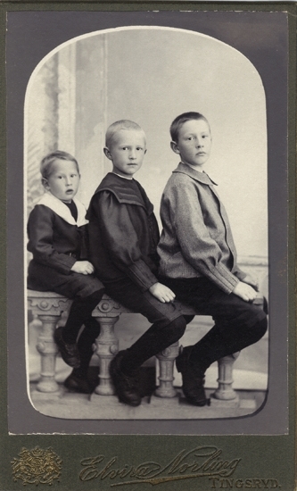 Grupporträtt (helfigur, halvprofil) av tre bröder (?), klädda i kortbyxor och bussaronger m.m. 
De sitter grensle på en barriär i en ateljé.