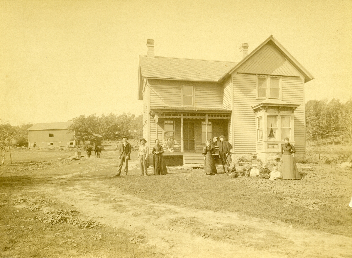 Flere kvinner, menn og barn er avbildet utenfor et hus på en farm i Amerika. I bakgrunnen er det en låve. Nr to fra venstre er Pål Milevatnet.