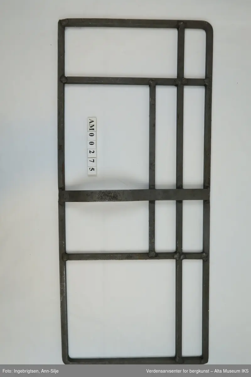 Rektangulær skifermål, med metallstenger smidd på tvers mellom både kortsidene og langsidene. På midten er det smidd på et håndtak.