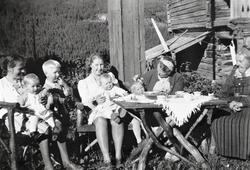 Gruppe i solveggen. Kvinner og barn. Kjole og sko.
Frå v.Ma