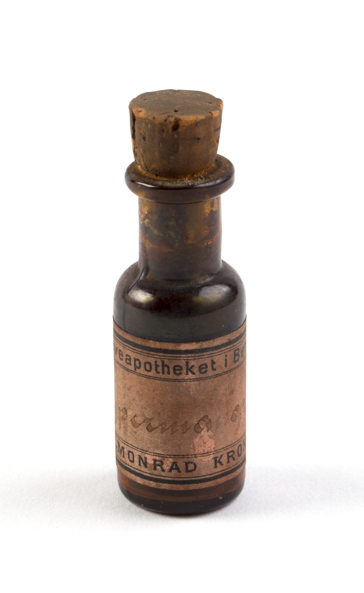 Brun flaske med brun kork, påført håndskrevet 
etikett, noe vanskelig å tyde.