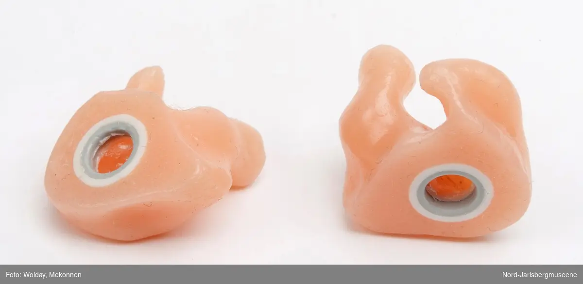 To ørepropper som kobles til høreapparat. Proppene er formet og tilpasset en spesiell bruker, og ligger i en liten plastpose med brukers navn.