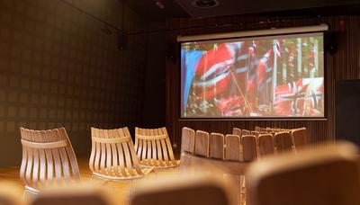 Bilde fra auditoriet i Wergelands Hus viser utsnitt av noen stoler vendt mot lerretet som viser et bilde fra 17.mai med norske flagg.