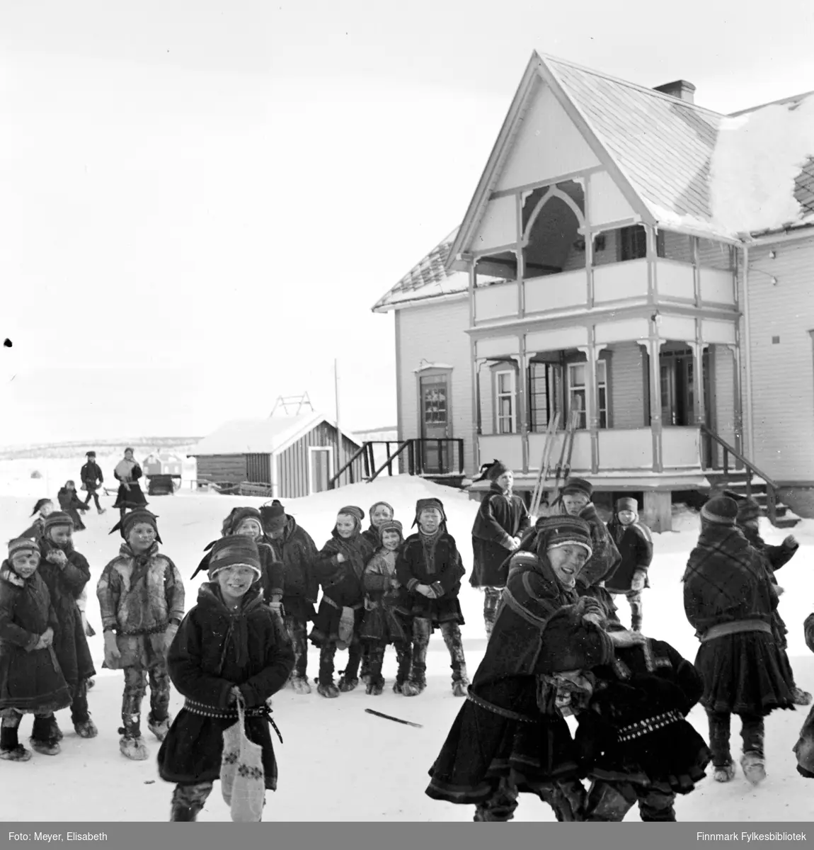 Internatstyrerboligen i Kautokeino, fotografert før krigen i perioden 1939-40. Samiske skolebarn leker i snøen utenfor. Til venstre i bildet bak ser man to voksne, muligens lærer og lærerinne. Alle barna er kledd i kofte eller pesk. I bakgrunnen står det også to par ski lent mot huset.