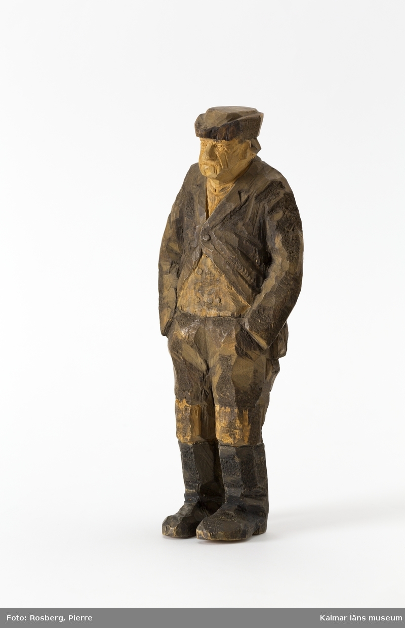 KLM 45932:2 Skulptur, av trä, målad. Figur i form av en stående man. Han har båda händerna i byxfickorna. Klädd i huvudbonad, jacka, väst och byxor med lappar på knäna, samt stövlar. Signerad på ryggen, H.C. Målad i bruna nyanser och svart. Tillverkad av Helge Rugland, f. Carlsson.