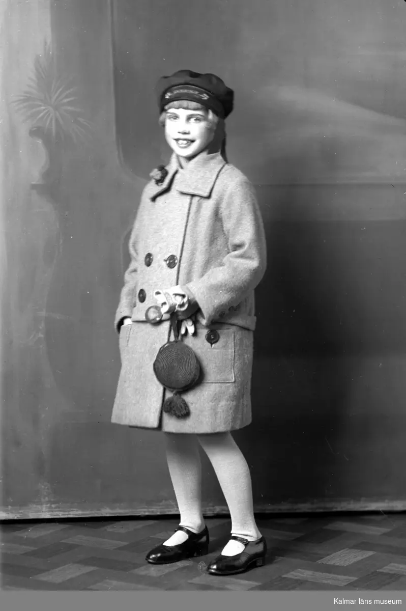 Ateljébild på en flicka med kappa och mössa. Enligt Walter Olsons journal är bilden beställd av Mary Holmquist.