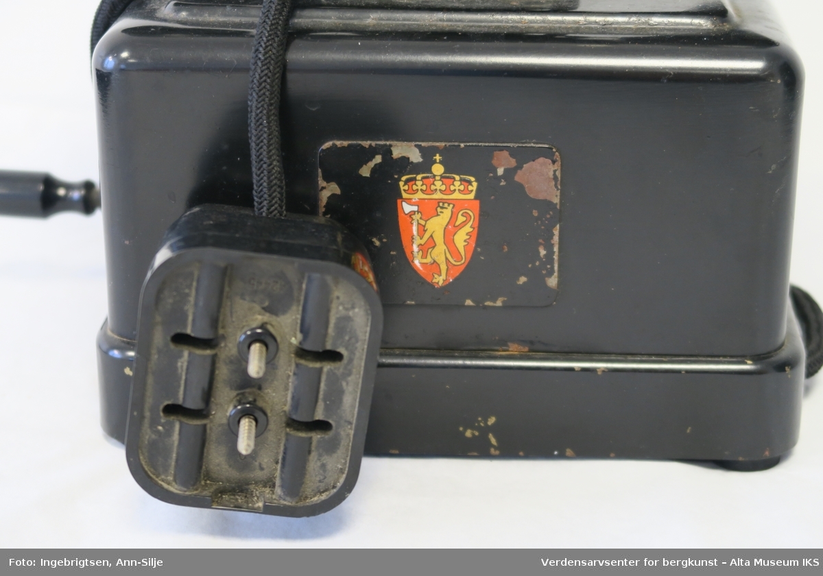 Prismeformet boks med bæreklo på toppen og telefonrør. Emblem med det norske riksvåpenet på framsiden av boksen. På siden er det festet en strømledning med støpsel i enden.