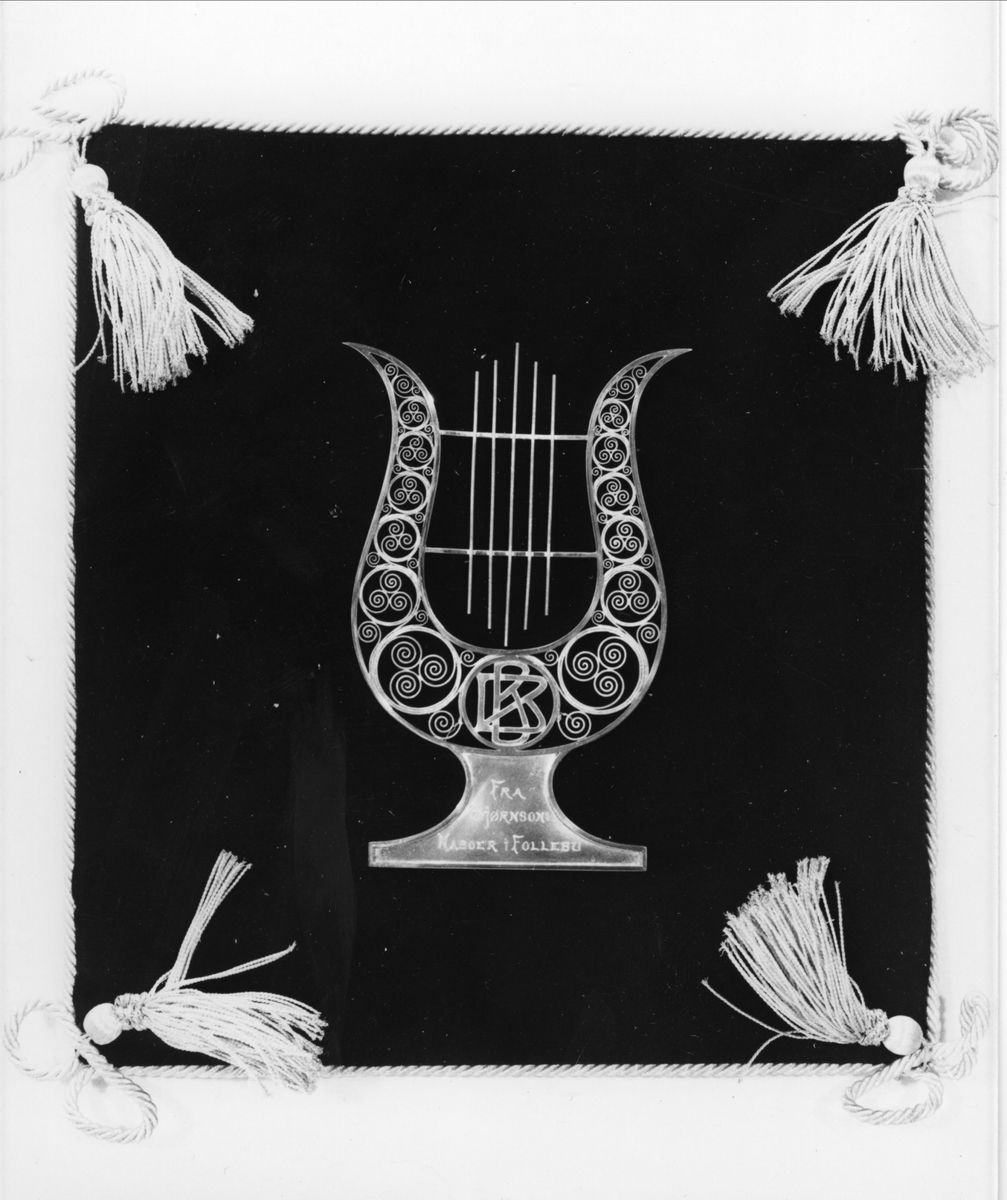 Lyre i sølv sydd fast på svart fløyelspute kantet med snorer og dusker i hjørnene. Initialene BB er utformet i jugendstil.