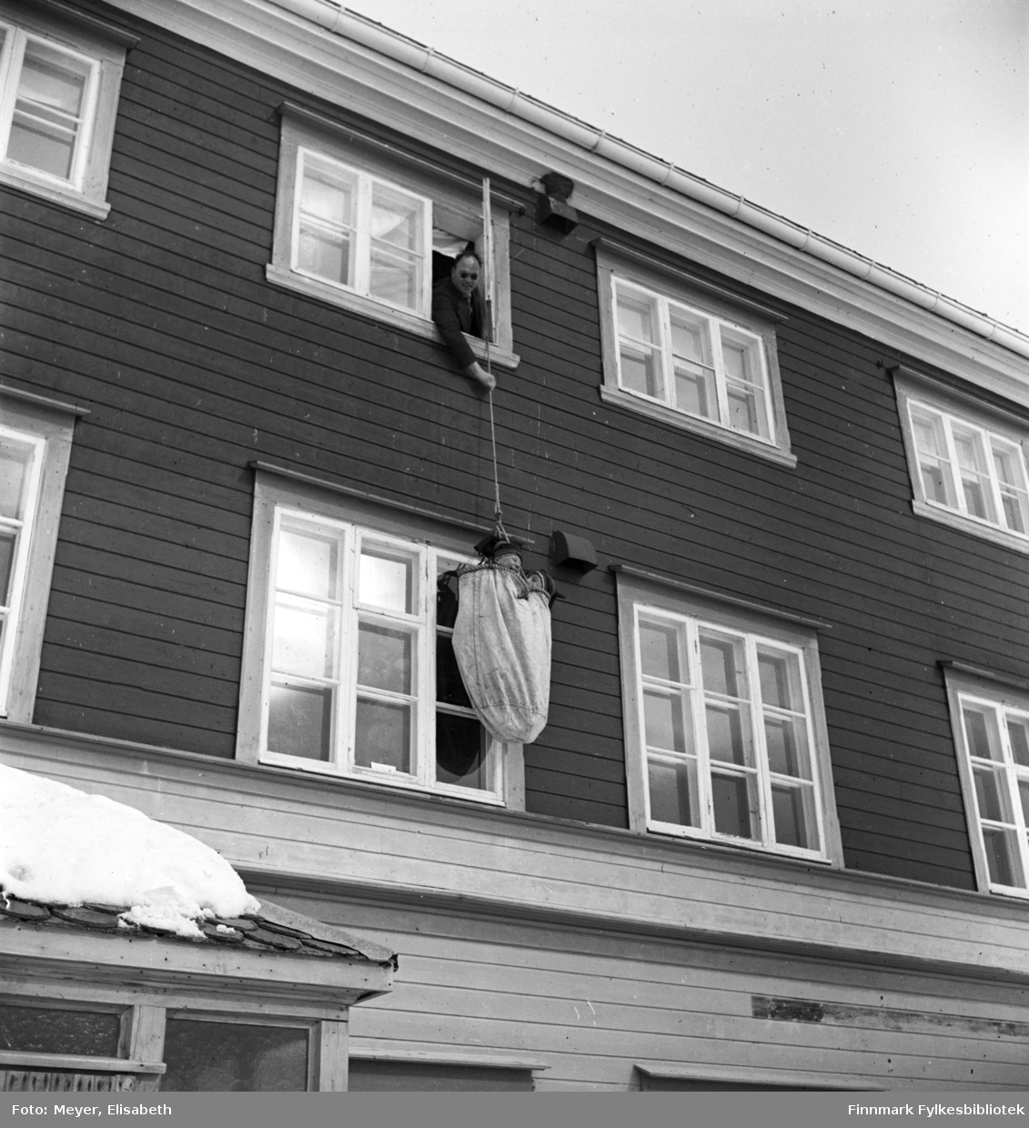 Brannøvelse på Kautokeino skoleinternat. Johan Nils Peder Johansen Sara og Johan Mikkelsen Sara henger på veggen oppi sekken festet til branntauet og blir firt ned 3 etasjer av en lærer med solbriller.