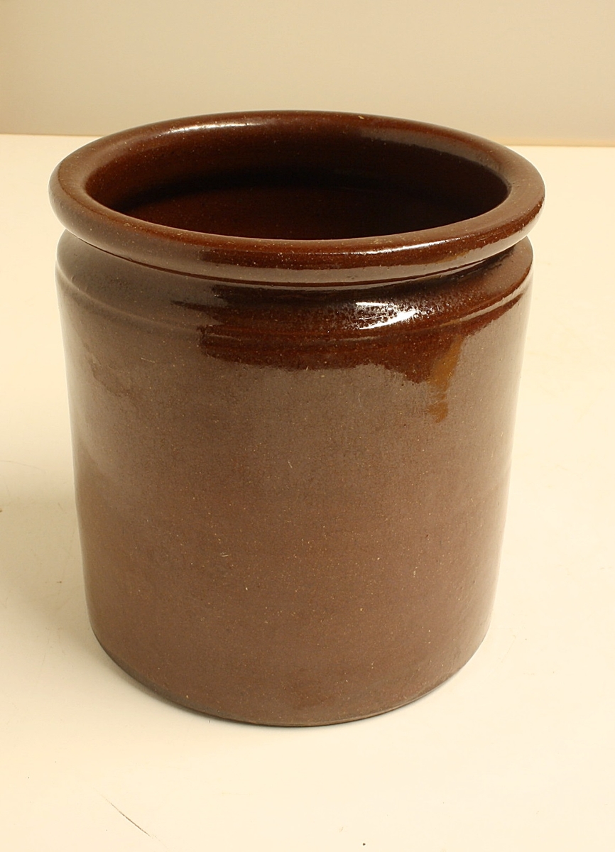 Sylinderformet sylterkukke med profilert munningsrand. Den er laget av brunglasert keramikk av rød leire.
