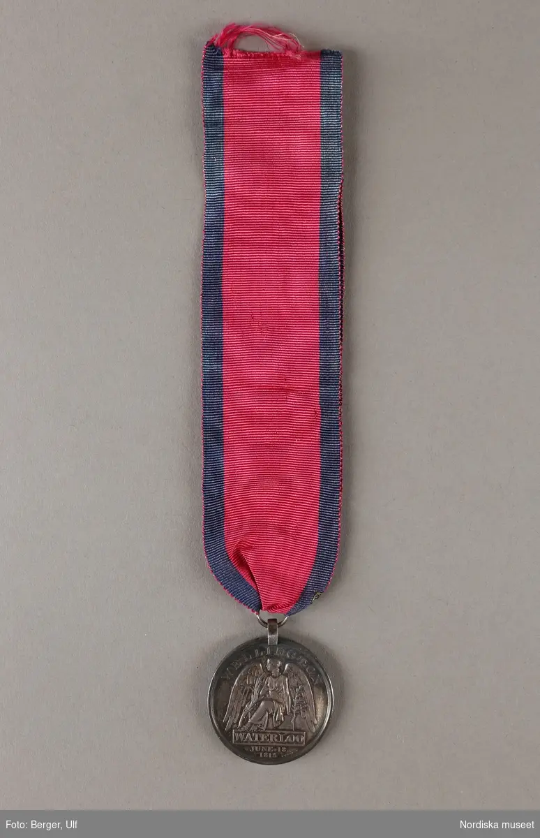 Huvudliggaren:
"Medalj. Se 34.443 [= 34.443-34.485 samlade af friherre Bennet.]
I tenn. Åtsidan: Georg IIIs bröstbild, v.s. och med omskrift: 'GEORGE P. REGENT'. Under bilden: 'T. WYON. JUN'. Frånsidan framställer segergudinnan, däröfver: 'WELLINGTON'. I afskärning: 'WATERLOO JUNE 18 1815'. Å kanten: 'JOHN SHEERMAN 2nd BATT. 59th REG:FOOT'. Diam 12 lin. 
I rödt band af sidenrips med blå ränder i kanterna.
Ink. ss. 34.424 [=ink. ss. 34.422  jämte 34.425-34.485]. [34.422=Ink. af byggmästare E. A. Boman i Stockholm 8/1 1883]"